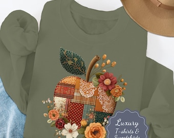 May include: An olive green sweatshirt featuring a patchwork apple design with floral accents. The apple is composed of various fabric patterns and textures, with a stem and leaf. The design includes red, orange, and white flowers. The text "Luxury T-shirts & Sweatshirts" is also visible.