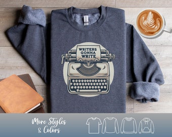 May include: A dark gray sweatshirt with a vintage typewriter graphic and the text "Writers Gonna Write." The design is set against a cream-colored circle. The image also shows a coffee cup and a stack of books.