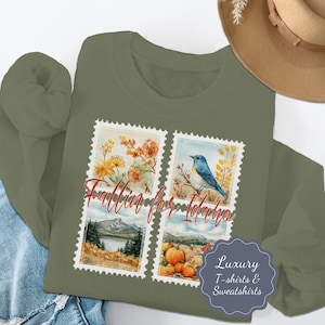 Puede incluir: Sudadera verde oliva con un diseño de cuatro paneles y el texto "Fallin' for Idaho". Los paneles muestran ilustraciones de acuarela de flores, un pájaro azul, un paisaje de montaña y calabazas. La sudadera también tiene una etiqueta "Luxury T-shirts & Sweatshirts".