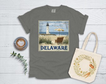 May include: A grey t-shirt featuring a vintage-style illustration of a lighthouse, beach, and umbrella. The text "DELAWARE" is printed below the image.