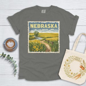 Op de afbeelding: Een donkergrijs T-shirt met een vintage-achtige illustratie van een veld in Nebraska. De illustratie is ingelijst als een postzegel en bevat de tekst "Nebraska".