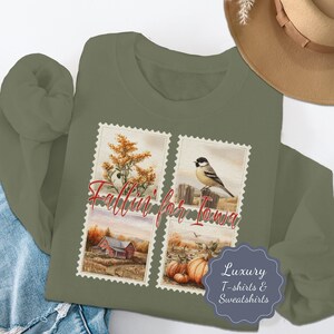 Puede incluir: Una sudadera verde oliva con un diseño de cuatro paneles que presenta ilustraciones en acuarela. Los paneles representan escenas de otoño: vara de oro, un pájaro en una valla, un granero rojo y calabazas. El texto "Fallin' for Iowa" está en escritura roja.