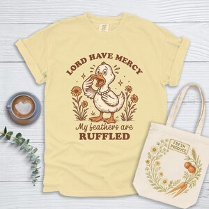 Könnte beinhalten: Hellgelbes T-Shirt mit einer Cartoon-Entengrafik, dem Text "LORD HAVE MERCY" und "My feathers are RUFFLED". Eine Tragetasche aus Segeltuch mit floralem Design und den Worten "FRESH PRODUCE" ist ebenfalls sichtbar.
