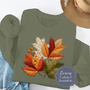 Op de afbeelding: Een olijfgroene sweatshirt met een applicatie van herfstbladeren in oranje, rood, geel en groen. De bladeren hebben gedetailleerde patronen en texturen. De tekst "Luxury T-shirts & Sweatshirts" staat in een blauw label.
