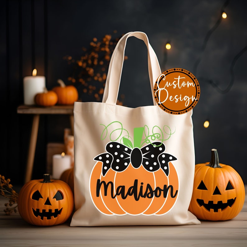 Op de afbeelding: Cr&egrave;me canvas draagtas met een oranje pompoenontwerp. De pompoen heeft een zwarte strik met witte stippen en de naam "Madison" in zwarte letters. Twee uitgesneden pompoenen flankeren de tas, wat een Halloween-thema cre&euml;ert.
