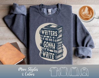 Puede incluir: Una sudadera gris oscuro con el texto "WRITERS GONNA WRITE" apilado sobre una pila de libros. El diseño es de color azul claro sobre un círculo color crema. La sudadera tiene cuello redondo y mangas largas.