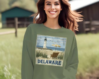May include: A green sweatshirt featuring a vintage-style graphic of a lighthouse on a beach with a beach chair and umbrella. The graphic is surrounded by a blue border and the word "DELAWARE" is printed below it.