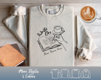May include: Cream-colored sweatshirt with a black graphic of a hand writing in a book, a coffee cup, and the words "Write On" and "Your Name Here." Additional shirt styles are shown at the bottom.