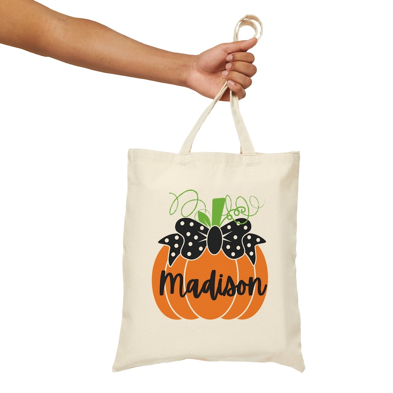 Op de afbeelding: Een cr&egrave;mekleurige draagtas met een oranje pompoenontwerp, een zwarte strik en de naam "Madison" in zwart schrift. De pompoen heeft groene ranken en een groene stengel. De tas wordt door een persoon vastgehouden.