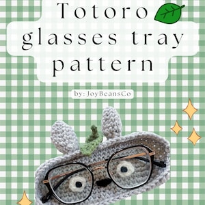 Może przedstawiać: Szydełkowa taca na okulary w kształcie Totoro, trzymająca parę okularów. Taca jest szara z uszami Totoro i zielonym liściem. Tło to zielono-biały wzór w kratę z napisem "Totoro glasses tray pattern".