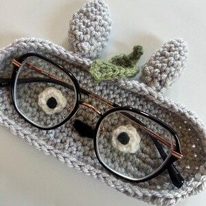 May include: A pair of black-framed eyeglasses rests in a handmade gray crochet glasses case. The case features a whimsical design with two gray ears and a green leaf, with a white and black flower detail.