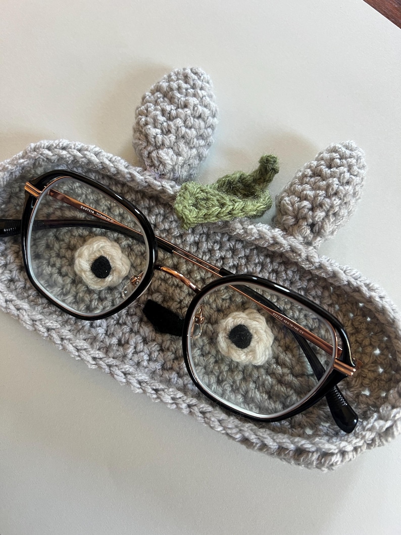 May include: A pair of black-framed eyeglasses rests in a handmade gray crochet glasses case. The case features a whimsical design with two gray ears and a green leaf, with a white and black flower detail.