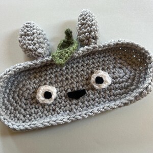 May include: A handmade, gray crochet tray with a whimsical face design. The tray features two gray, cone-shaped ears, white and black eyes, a black mouth, and a small green leaf accent. The tray is perfect for holding small items.