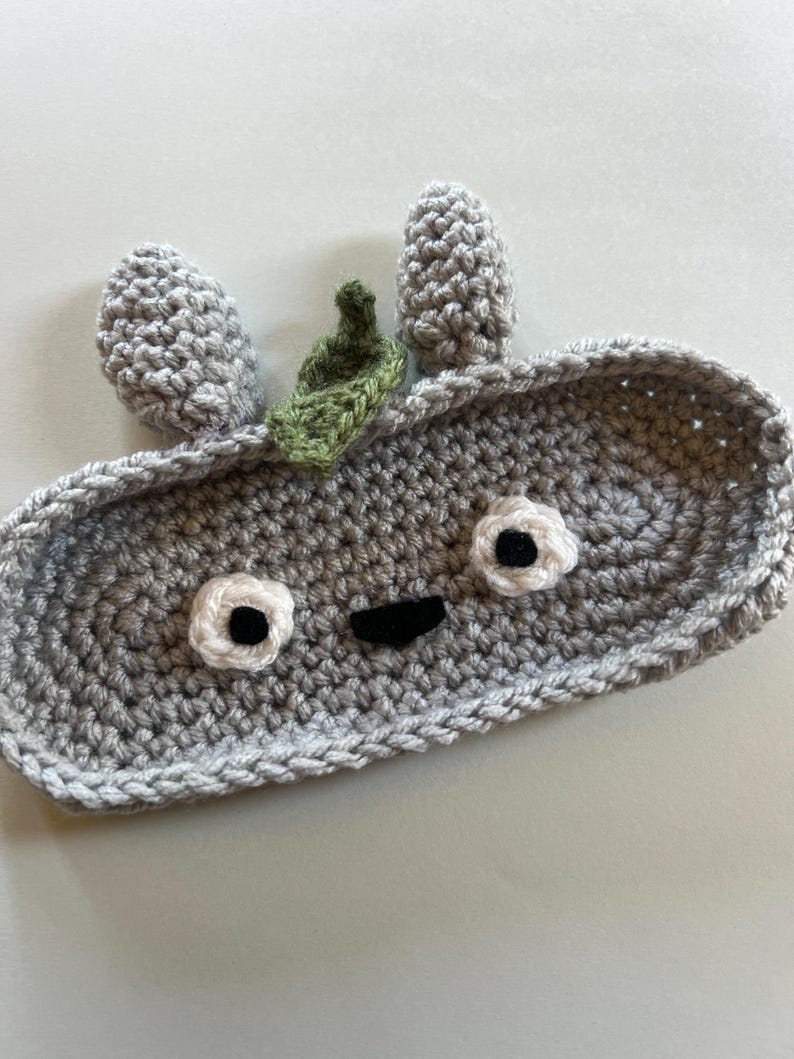May include: A handmade, gray crochet tray with a whimsical face design. The tray features two gray, cone-shaped ears, white and black eyes, a black mouth, and a small green leaf accent. The tray is perfect for holding small items.