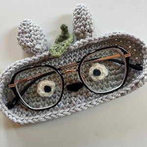 May include: A gray crocheted glasses case with a whimsical design, featuring two gray ears and a green leaf. The case holds a pair of round eyeglasses with black frames and gold-colored arms. The case has a face design with white and black accents.