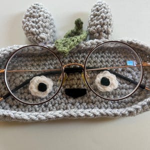 May include: Round eyeglasses with a thin, gold-colored frame rest on a gray crocheted eyeglass case. The case is shaped like a creature with white and black flower-shaped eyes, a black nose, and gray ears and a green leaf.