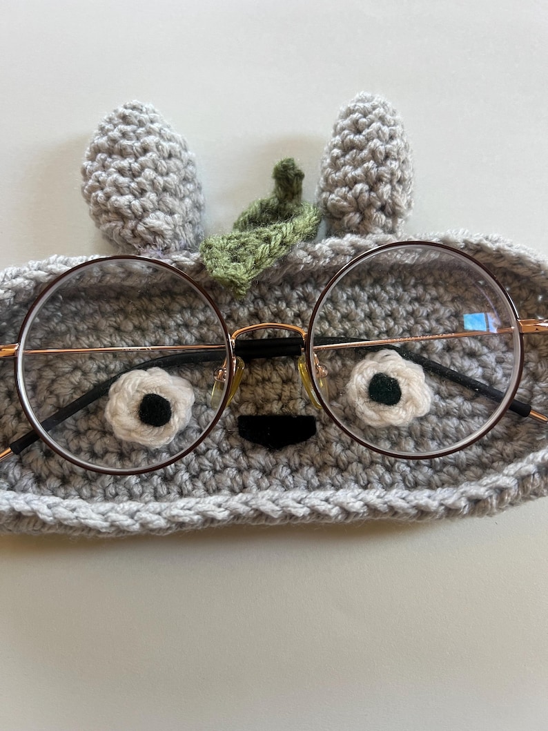 May include: Round eyeglasses with a thin, gold-colored frame rest on a gray crocheted eyeglass case. The case is shaped like a creature with white and black flower-shaped eyes, a black nose, and gray ears and a green leaf.