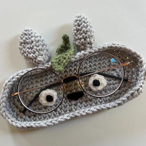 May include: A gray crocheted glasses holder shaped like a face with round glasses. The holder has two gray ears and a green leaf. The glasses have a gold frame and black arms.