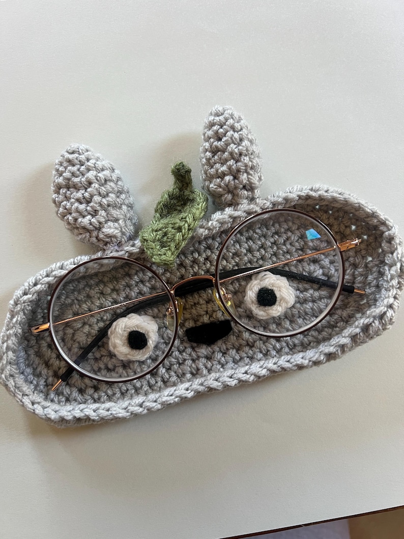 May include: A gray crocheted glasses holder shaped like a face with round glasses. The holder has two gray ears and a green leaf. The glasses have a gold frame and black arms.