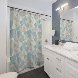 May include: A shower curtain featuring an abstract pattern of overlapping organic shapes in shades of soft blue, sage green, and beige. The curtain hangs from a black rod with metal rings, displaying a watercolor-inspired design across the fabric.