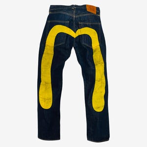 Puede incluir: Vaqueros de mezclilla azul índigo oscuro con un gran gráfico de gaviota amarillo pintado a mano en los bolsillos traseros y las piernas. Los vaqueros incluyen una etiqueta de cuero marrón en la cintura trasera con texto en relieve y una etiqueta roja.