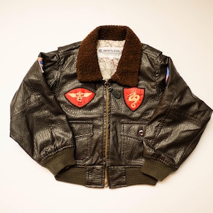 May include: A dark brown faux leather bomber jacket featuring a brown sherpa collar, a front zipper closure, and two front flap pockets. The chest displays two red patches with gold embroidery, and the interior label reads HOUSTON.