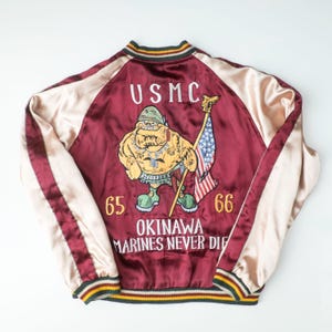 May include: A maroon and cream satin souvenir jacket featuring a back embroidery of a cartoon character holding a US flag, the text USMC, 65, 66, OKINAWA, and MARINES NEVER DIE. The jacket has striped ribbed cuffs, collar, and hem in maroon, cream, and green.
