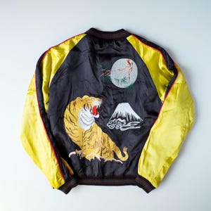 May include: A black and yellow satin bomber jacket featuring detailed embroidery on the back. The design includes a large yellow tiger, a white mountain peak, and a circular moon with pine branches, all rendered in vibrant thread colors.