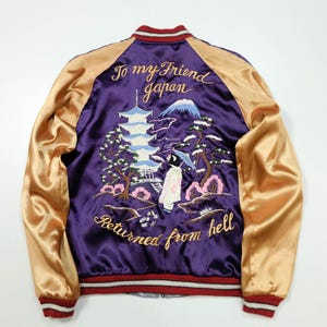 Puede incluir: Una chaqueta bomber de satén morado con mangas doradas y ribetes de punto a rayas. La espalda presenta un bordado intrincado de una pagoda, una montaña, un pino y una figura con paraguas, con el texto "To my Friend japan Returned from hell."