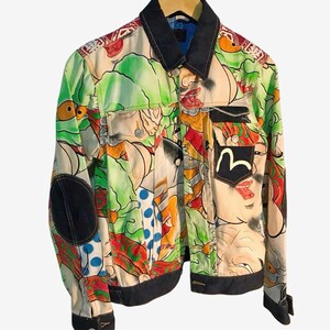 Puede incluir: Una chaqueta de manga larga con un estampado gráfico multicolor de rostros estilizados y formas abstractas. La prenda incluye cuello, puños, dobladillo y coderas en color oscuro, con un bolsillo en el pecho que muestra un logotipo blanco.