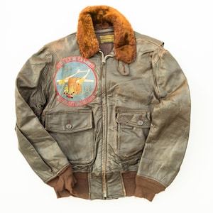 Puede incluir: Una chaqueta de vuelo de cuero marrón con cuello de piel marrón oscuro, cremallera frontal y dos bolsillos con solapa y botón. Un parche circular en el pecho presenta una ilustración de un avión con el texto FAR EAST 60 y 253 HEAVY RIDERS.