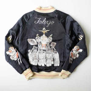 May include: A black satin souvenir jacket featuring cream-colored ribbed cuffs and hem with red and yellow stripes. The back displays intricate embroidery of people carrying a portable shrine, the word Tokyo, and figures on ladders on the sleeves.