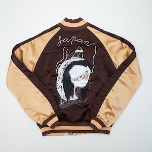 Puede incluir: Una chaqueta bomber de satén marrón y beige con un bordado en la espalda de una linterna blanca estilizada con un rostro y cabello negro largo. La palabra "japan" está bordada en letra cursiva blanca sobre la linterna. Puños y cuello acanalados.