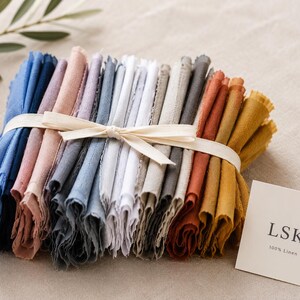 Può includere: Un fascio di tovaglioli di lino piegati in vari colori, tra cui blu, rosa, grigio e giallo, legati con un nastro color crema. Un piccolo biglietto con il testo "LSK 100% Linen" è posto accanto ai tovaglioli.
