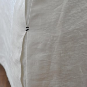 Può includere: Primo piano di una gonna da letto in lino bianco. Il tessuto ha un aspetto strutturato con una cucitura visibile e cuciture decorative in filo scuro. L'angolo della gonna è visibile, appoggiato su un pavimento in legno.
