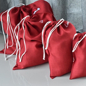 Pode incluir: Uma coleção de sacos vermelhos com cordão e cordões brancos. Alguns sacos têm detalhes bordados, incluindo um gorro de Pai Natal e o nome "Emi". Os sacos estão dispostos em fila, de tamanhos variados.