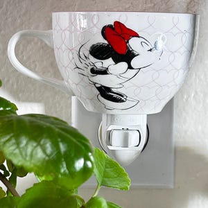 May include: A white ceramic wall plug-in night light shaped like a teacup. The cup features a repeating light pink heart pattern and a black and white illustration of Minnie Mouse wearing a bright red bow. The base has a white toggle switch.