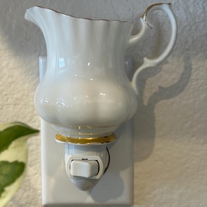 Puede incluir: Una luz de noche de porcelana blanca con forma de jarrita. La jarra tiene un borde festoneado y un ribete dorado. La base de la luz de noche tiene un anillo dorado y un interruptor. Está enchufada a una toma de corriente blanca.