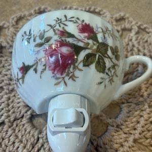 May include: White porcelain night light shaped like a teacup, adorned with pink roses and green leaves. The night light has a handle and a white plastic base with an on/off switch. The teacup is decorated with gold accents.