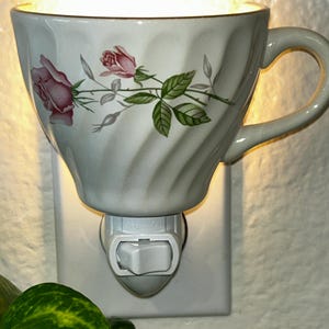 Puede incluir: Luz de noche con forma de taza de té blanca con un diseño floral de rosas rosas. La taza de té tiene un asa y está montada en una base blanca enchufable con un interruptor de encendido/apagado. La taza se ilumina desde dentro.