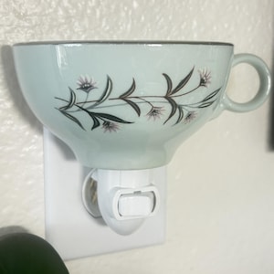 Puede incluir: Una luz nocturna con forma de taza de té azul claro con un diseño floral. La taza tiene un asa y está montada sobre una base de luz nocturna blanca. El diseño floral presenta tallos marrones oscuros y flores moradas.