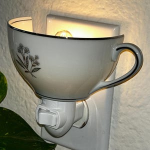 Peut inclure: Veilleuse en forme de tasse à thé blanche avec une bordure noire et un motif floral. La veilleuse est branchée sur une prise murale blanche et possède un interrupteur marche/arrêt. L'ampoule est visible à l'intérieur de la tasse.