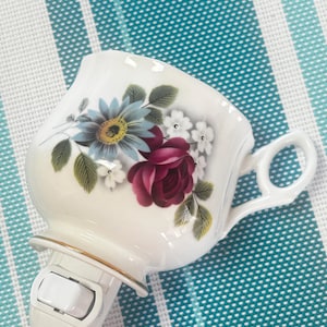 Puede incluir: Una luz de noche con forma de taza de té de porcelana blanca, adornada con un diseño floral con flores azules, rojas y blancas. Tiene un interruptor de encendido/apagado blanco y un borde dorado. El fondo es un patrón de rayas turquesas y blancas.