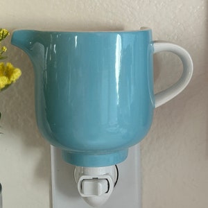 Puede incluir: Un calentador de cera eléctrico de cerámica azul claro con forma de jarra, con asa blanca y un interruptor de encendido/apagado.