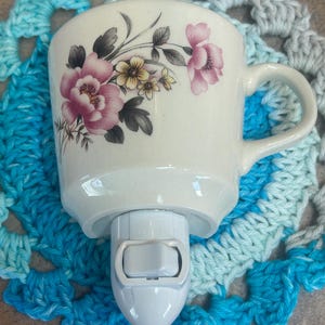 Puede incluir: Una luz de noche de cerámica blanca con forma de taza, adornada con diseños florales rosas y amarillos. La luz de noche tiene un interruptor de encendido/apagado y está colocada sobre un posavasos de ganchillo azul y blanco. La taza tiene un asa.