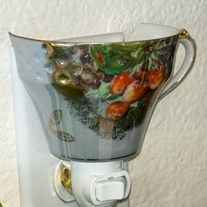 Puede incluir: Una luz de noche decorativa con un diseño de taza de té de porcelana. La taza de té tiene un borde y un asa dorados, con un colorido patrón de frutas y hojas. La luz de noche es blanca y tiene un interruptor de encendido/apagado.