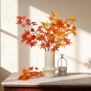 May include: A white vase holds vibrant faux maple branches with red, orange, and yellow leaves. A small bundle of leaves rests on the table. A candle in a wire holder with the word "House" is also on the table.