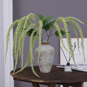 May include: A decorative arrangement featuring cascading green, flowering stems and green leaves in a speckled, beige ceramic vase. The vase sits on a dark brown wooden table, with a diffuser and framed art in the background.