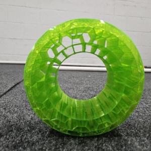 May include: A bright green, 3D printed, hollow, cylindrical object with a honeycomb pattern. The object is lying on a gray carpet.