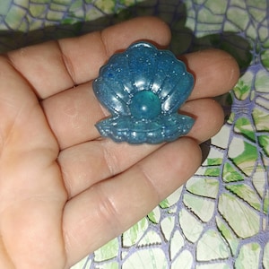 Può includere: Un guscio di conchiglia blu glitterato con una perla verde all'interno. Il guscio è fatto di resina ed è un oggetto decorativo.